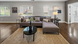 A modern living room with a sectional sofa, a coffee table, and a rug. There are two side tables with lamps, a framed picture on the wall, and a door to the right.