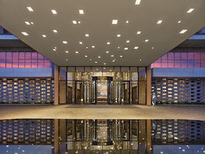 Een moderne gevel met glazen wanden, een reflecterend waterkunstwerk ervoor en een reeks verlichtingen in het plafond.