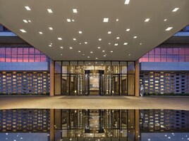 Una entrada de edificio moderna con fachada de vidrio, una fuente de agua reflectante en primer plano y una serie de luces en el techo.