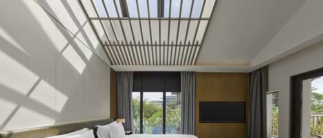 A modern hotel room with a large bed, a flat-screen TV, and a view of greenery outside.