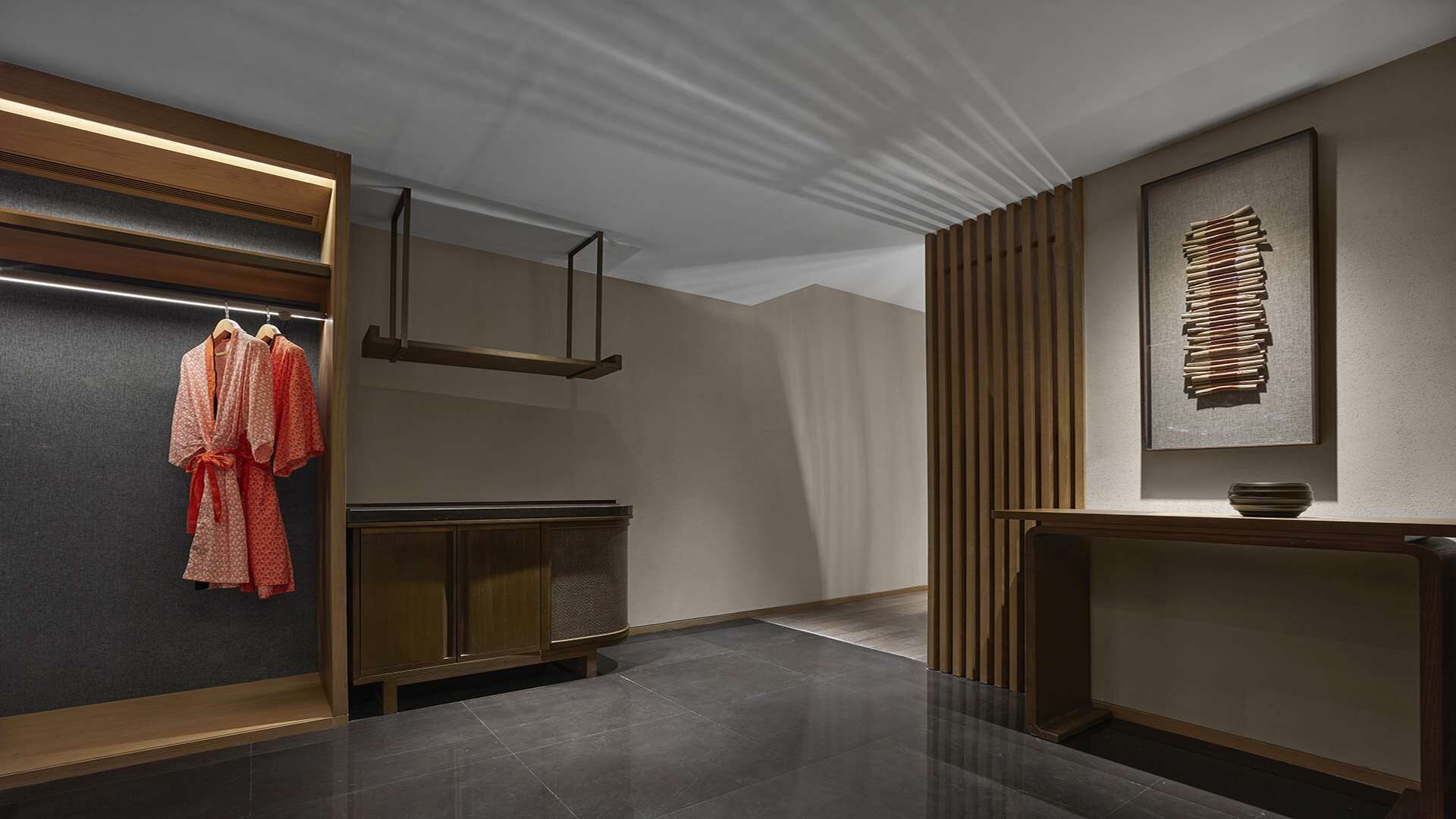 A modern hotel lobby with a dark tiled floor, a wooden reception desk, a wall-mounted TV, and a clothing rack with red robes.