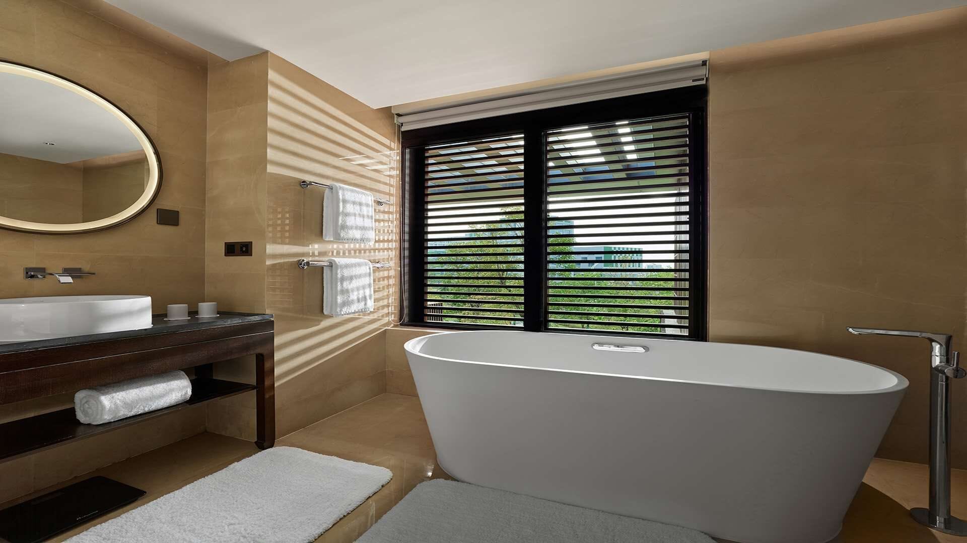 A modern bathroom with a freestanding bathtub, a large mirror, and a window with blinds.
