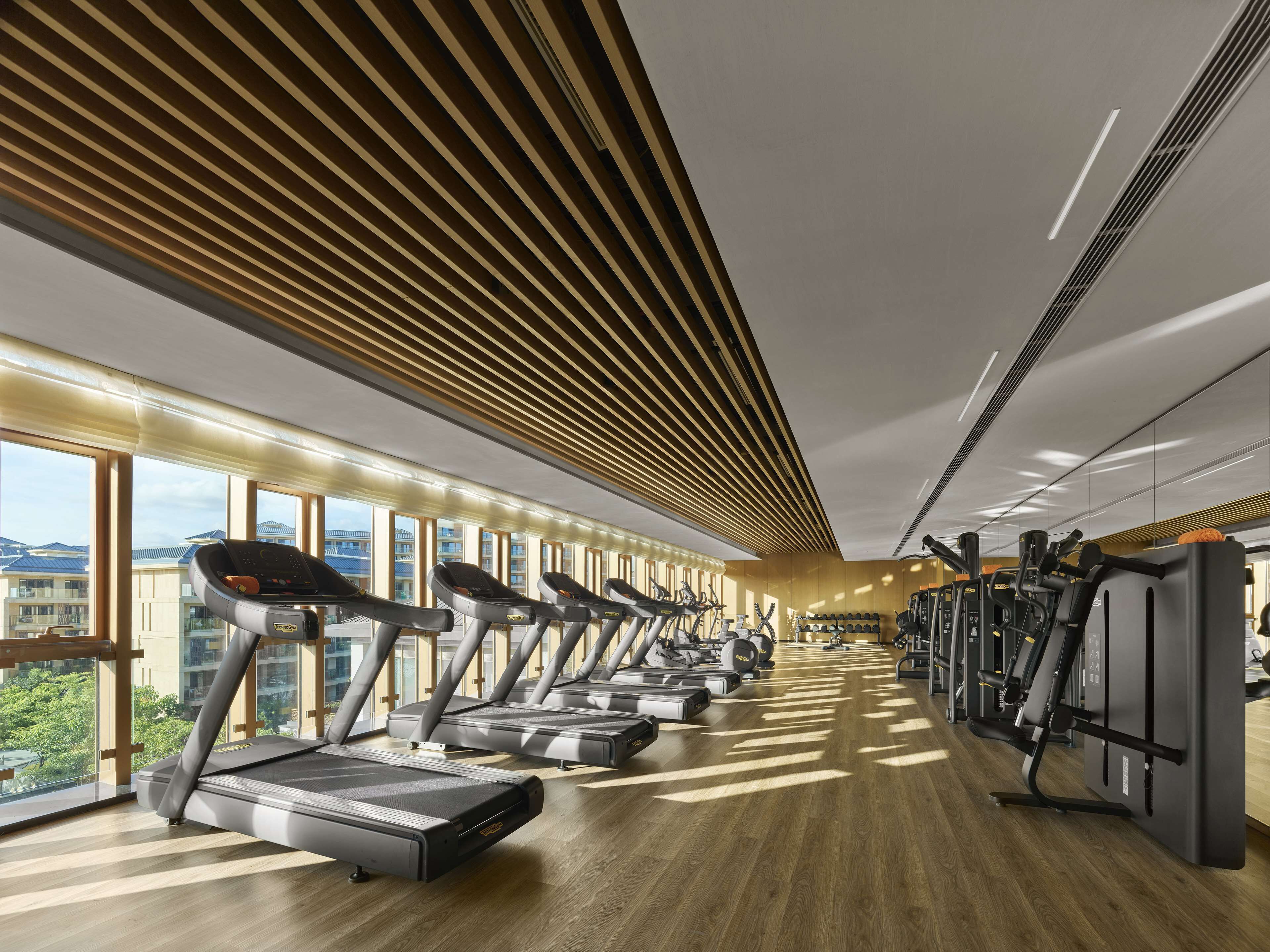A modern gym with treadmills, elliptical machines, and weight equipment. Large windows offer views of buildings and trees.