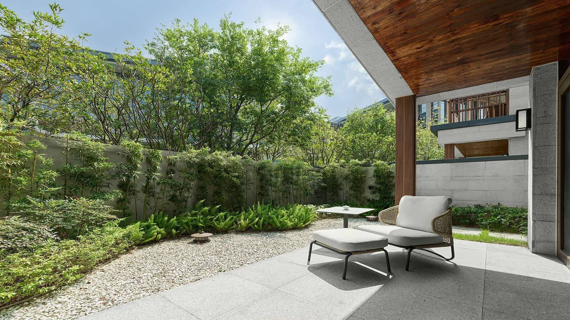 A modern outdoor patio with a stone table and two chairs, surrounded by lush greenery and a stone wall.