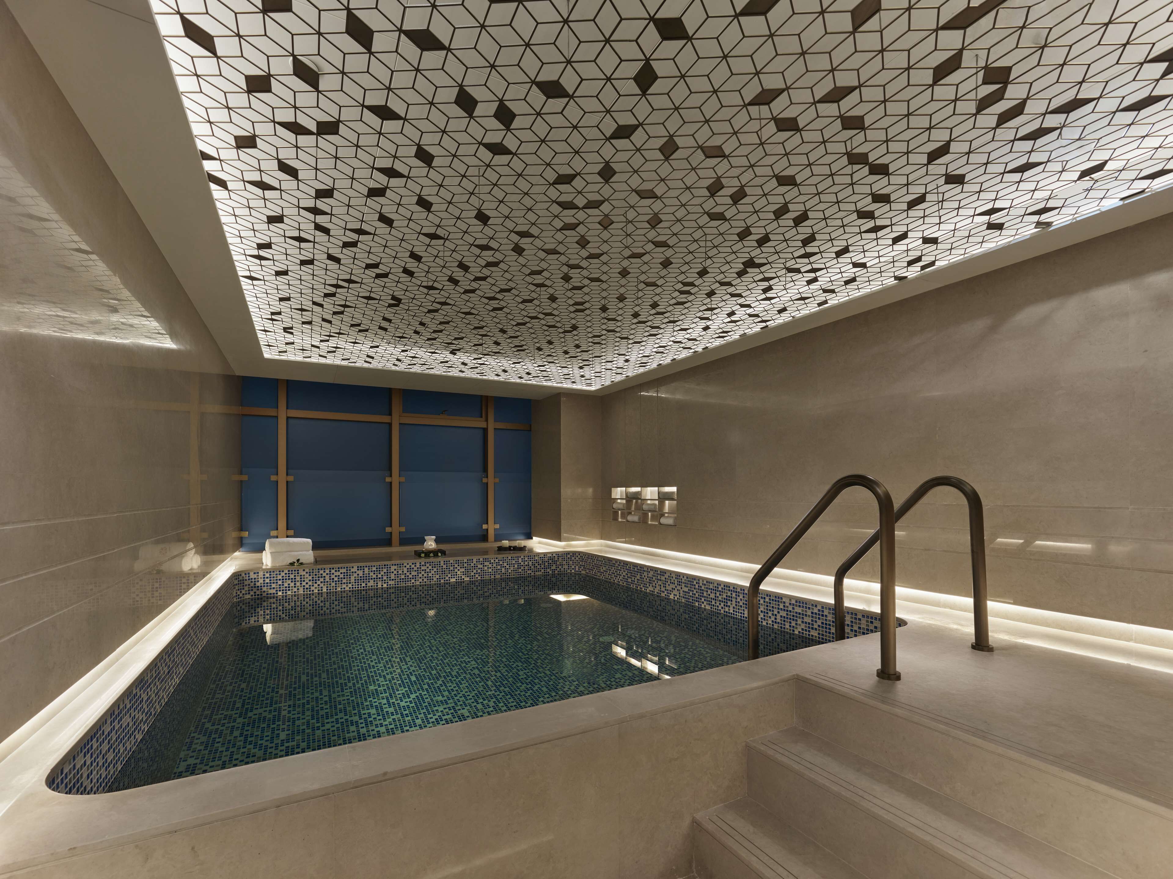 A modern indoor swimming pool with geometric ceiling tiles, a blue-tiled wall, and a set of stairs leading to a jacuzzi area.