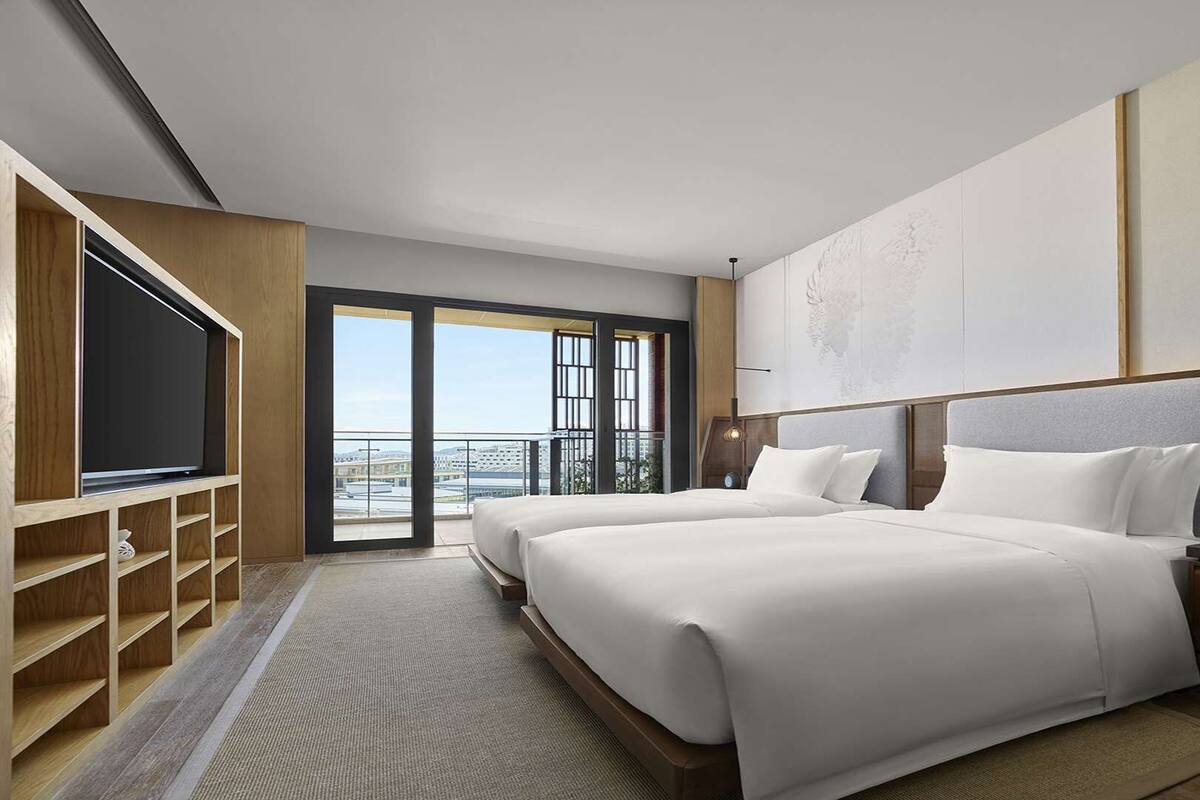 Une chambre d’hôtel avec deux lits, une télévision, un bureau avec une lampe et une grande fenêtre donnant sur un balcon.