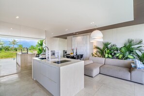 Una cocina moderna con una isla central, un área de comedor con un sofá y vista a un paisaje tropical.