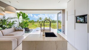 A modern kitchen with a large island, a sofa, and a view of a lush garden. - Green Flow Villa 2 with Private Pool in Ubud (Ubud)