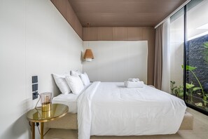A modern hotel room with a large bed, a bedside table, a lamp, and a view of greenery outside.