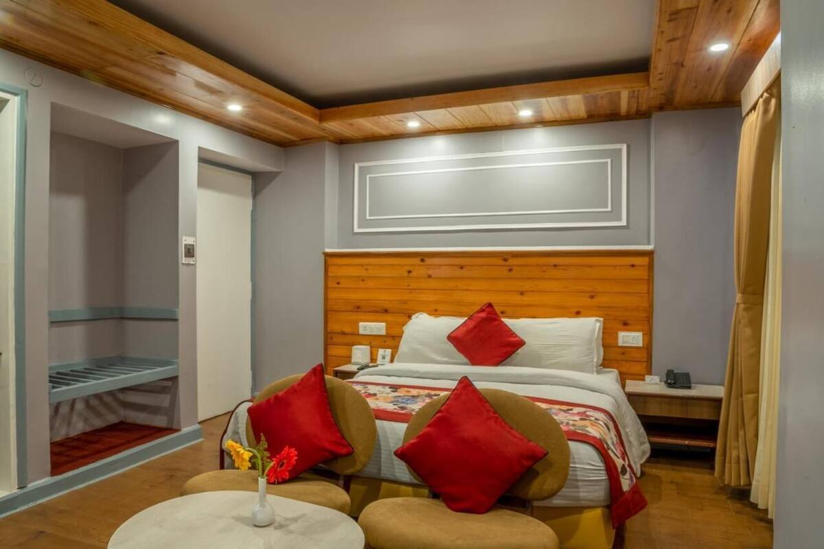 A bedroom with a wooden headboard, a bed with red pillows, a bedside table, and a chair with a vase of flowers.