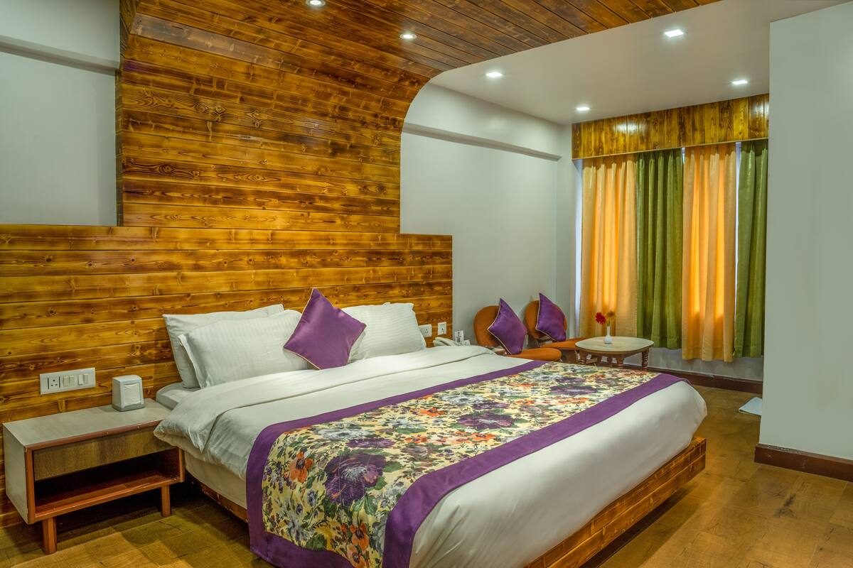 A bedroom with a wooden headboard, a bed with white bedding and purple pillows, a nightstand with a lamp, and a window with curtains.