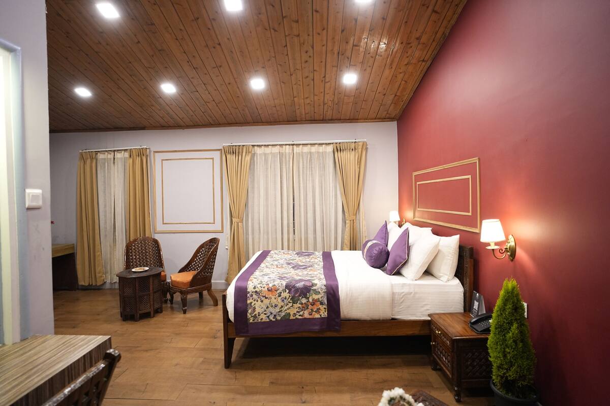 A bedroom with a wooden ceiling, a bed with a floral bedspread, two chairs, a small table, and a nightstand with a lamp.