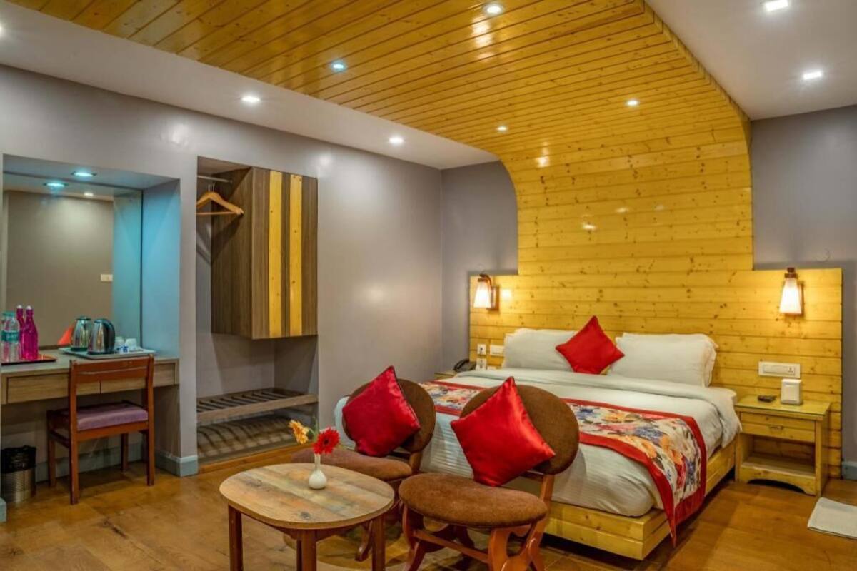 A hotel room with a wooden ceiling, a bed with red pillows, a desk with a chair, and a small table with a vase.