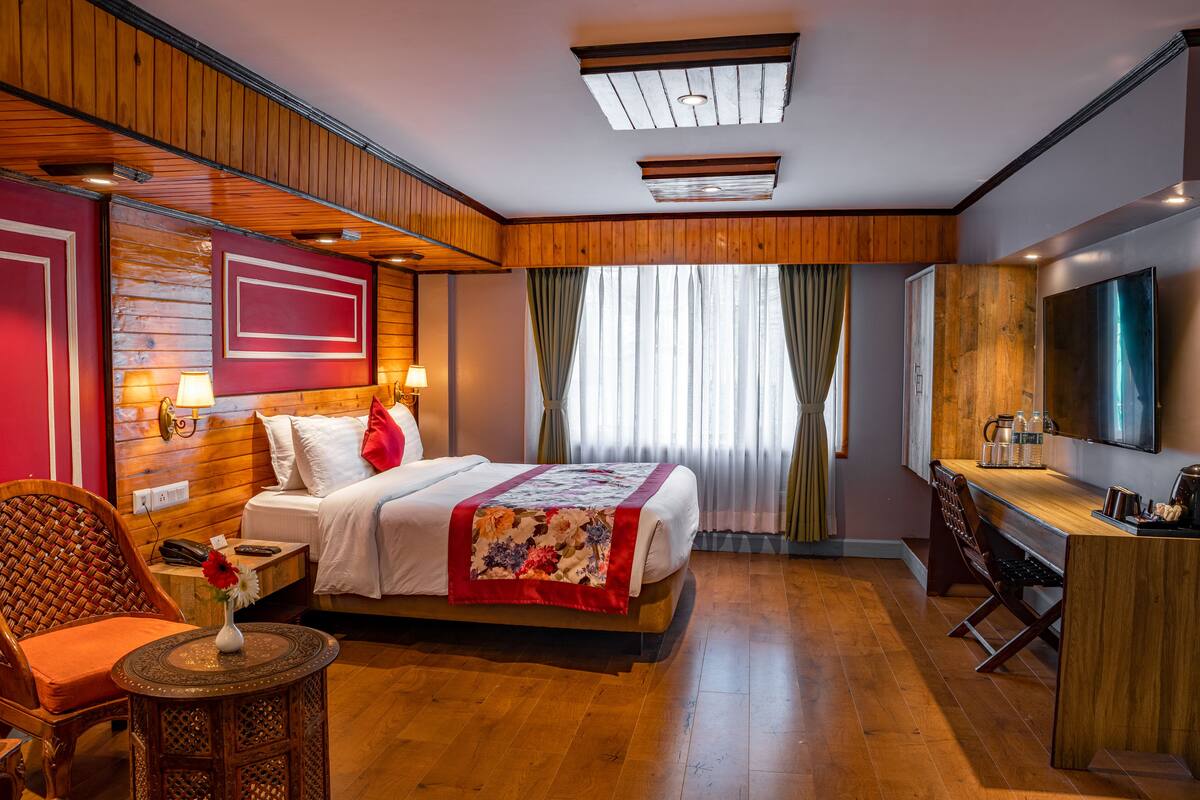 A hotel room with a wooden floor, a bed with a red and white bedspread, a desk with a television, and a window with curtains.