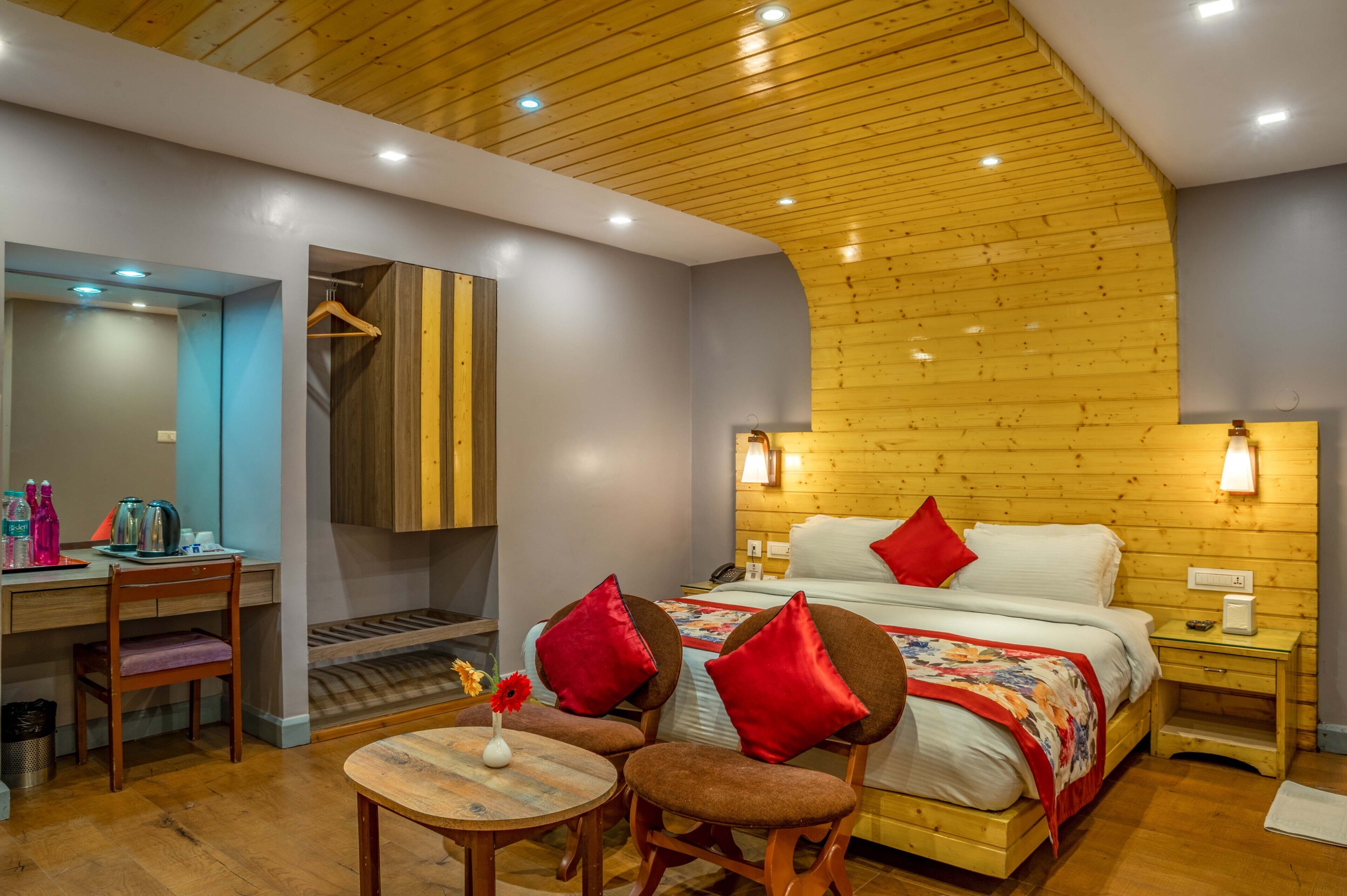 A cozy bedroom with a wooden ceiling, a bed with red pillows, a bedside table, and a desk with a chair.