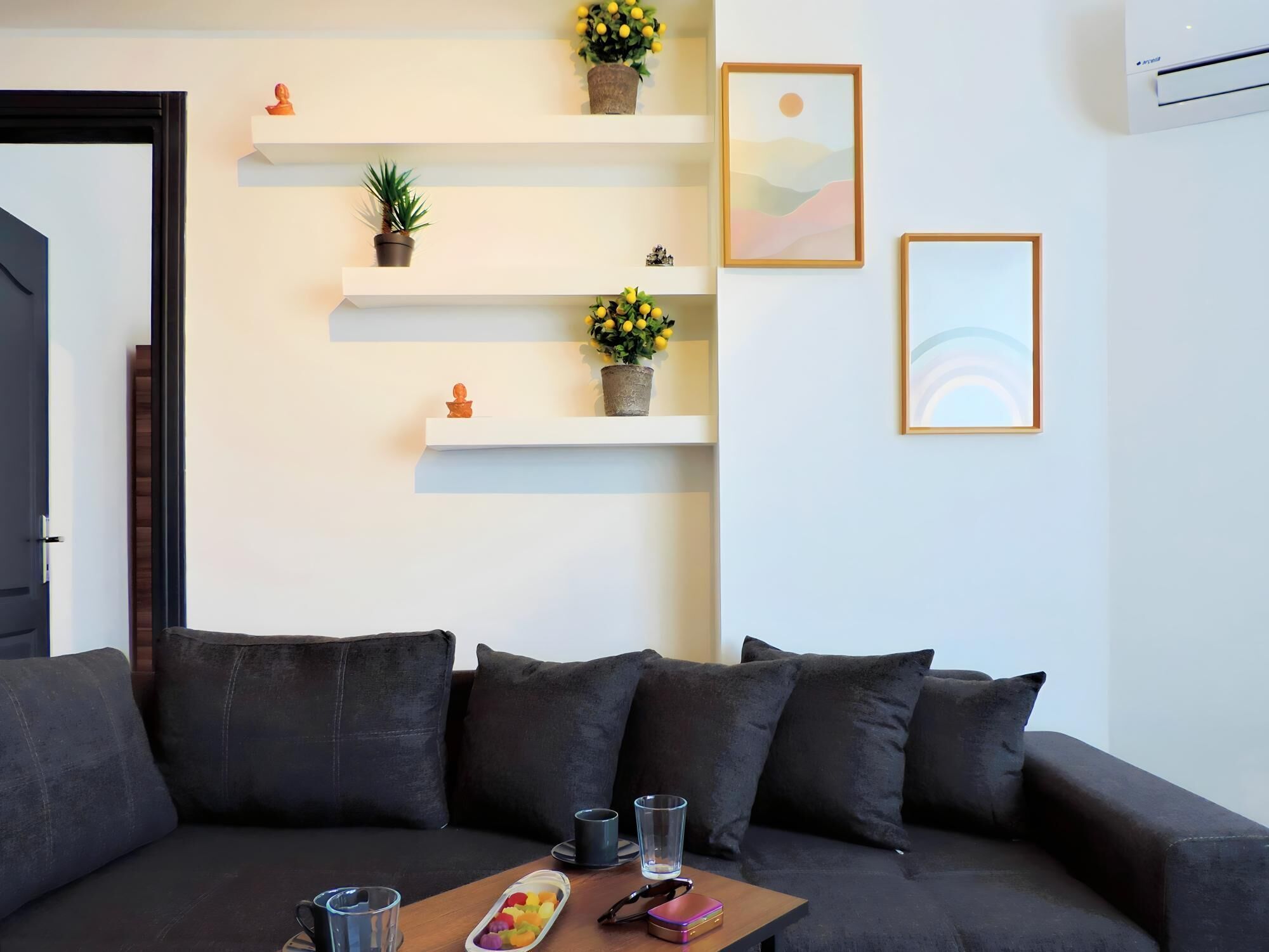 A modern living room with a dark grey sofa, a wooden coffee table with a tray of colorful objects, and white shelves with decorative items and plants.