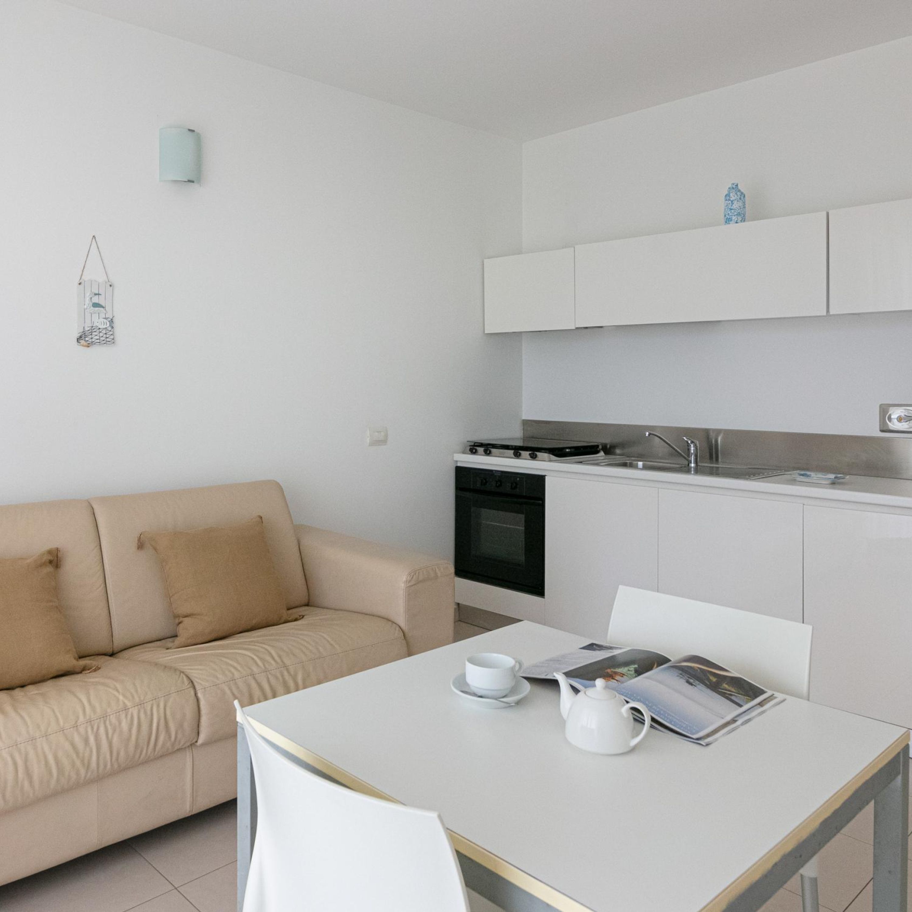 A modern kitchen with white cabinets, a stove, and a sink. There is a beige sofa with two pillows and a white table with a teapot and cups.