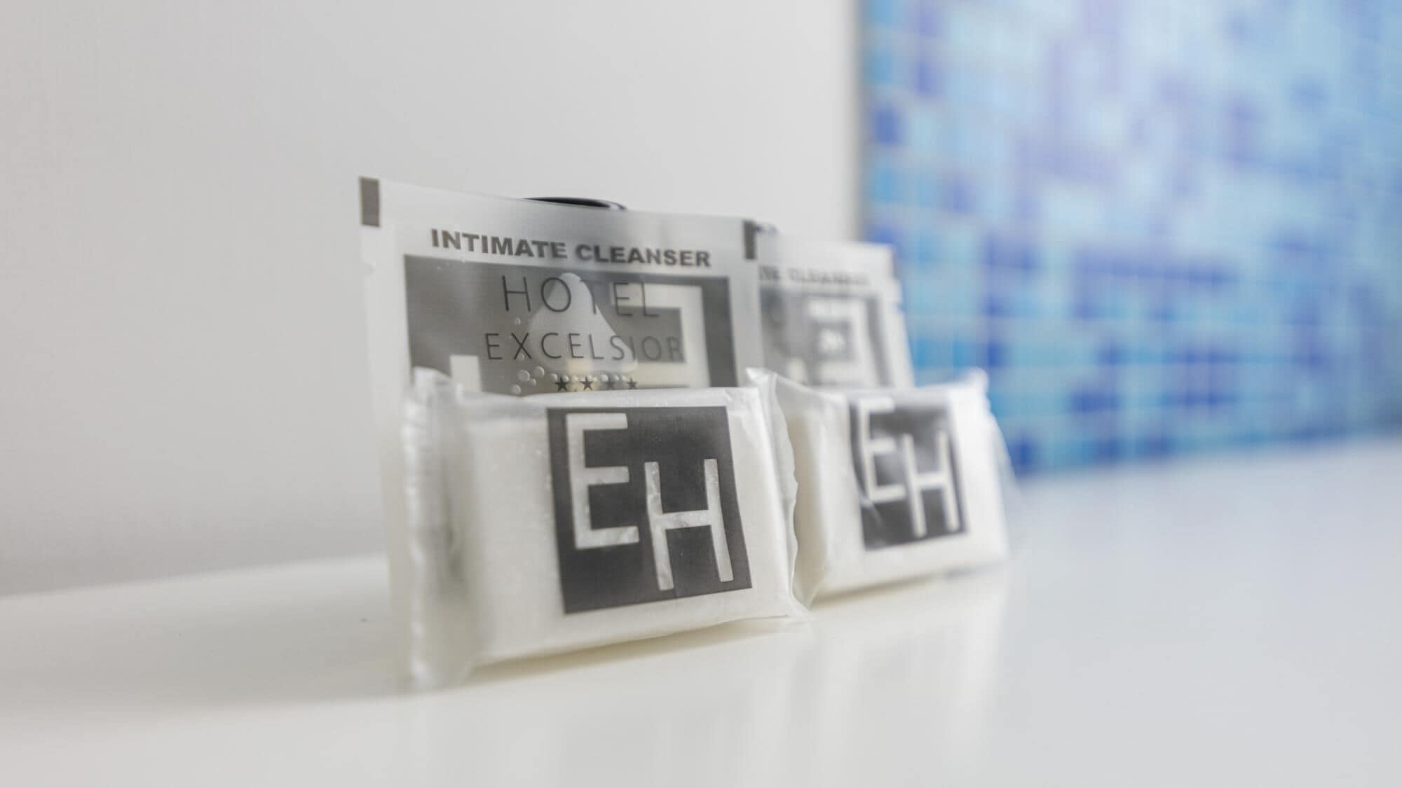 A hotel room with a white counter displaying a minibar with a minibar key card holder labeled "INTIMATE CLEANSER" and "HOTEL EXCELSIOR."
