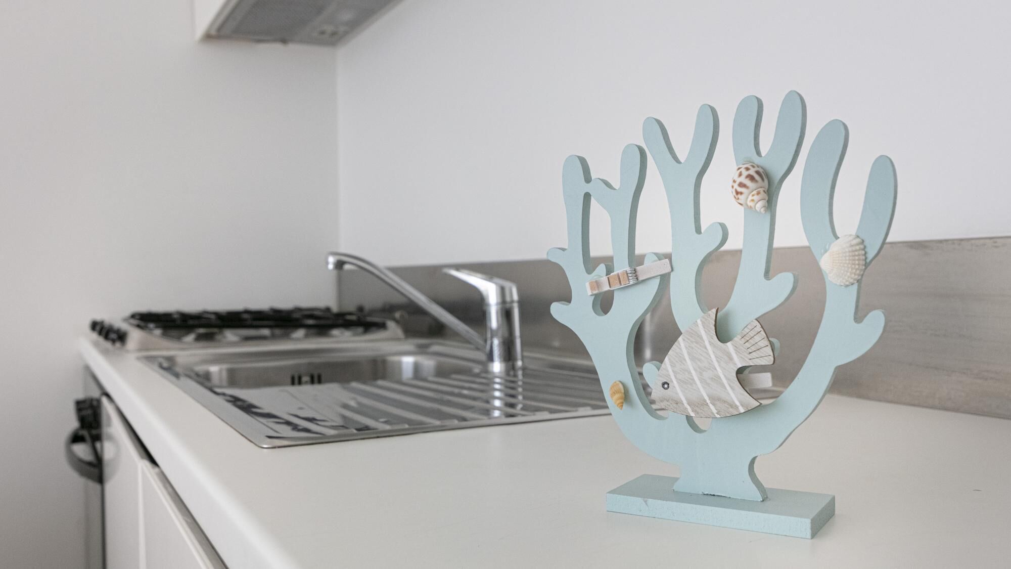 Una cocina con encimera blanca, pileta de acero inoxidable y un adorno azul de coral con conchas marinas.