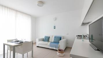 A modern, minimalist living room with a white sofa, a dining table with chairs, and a kitchen area with a large TV.