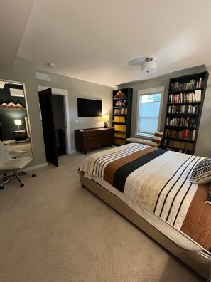 Um quarto com uma cama grande, uma escrivaninha com cadeira, uma estante de livros e uma TV na parede.