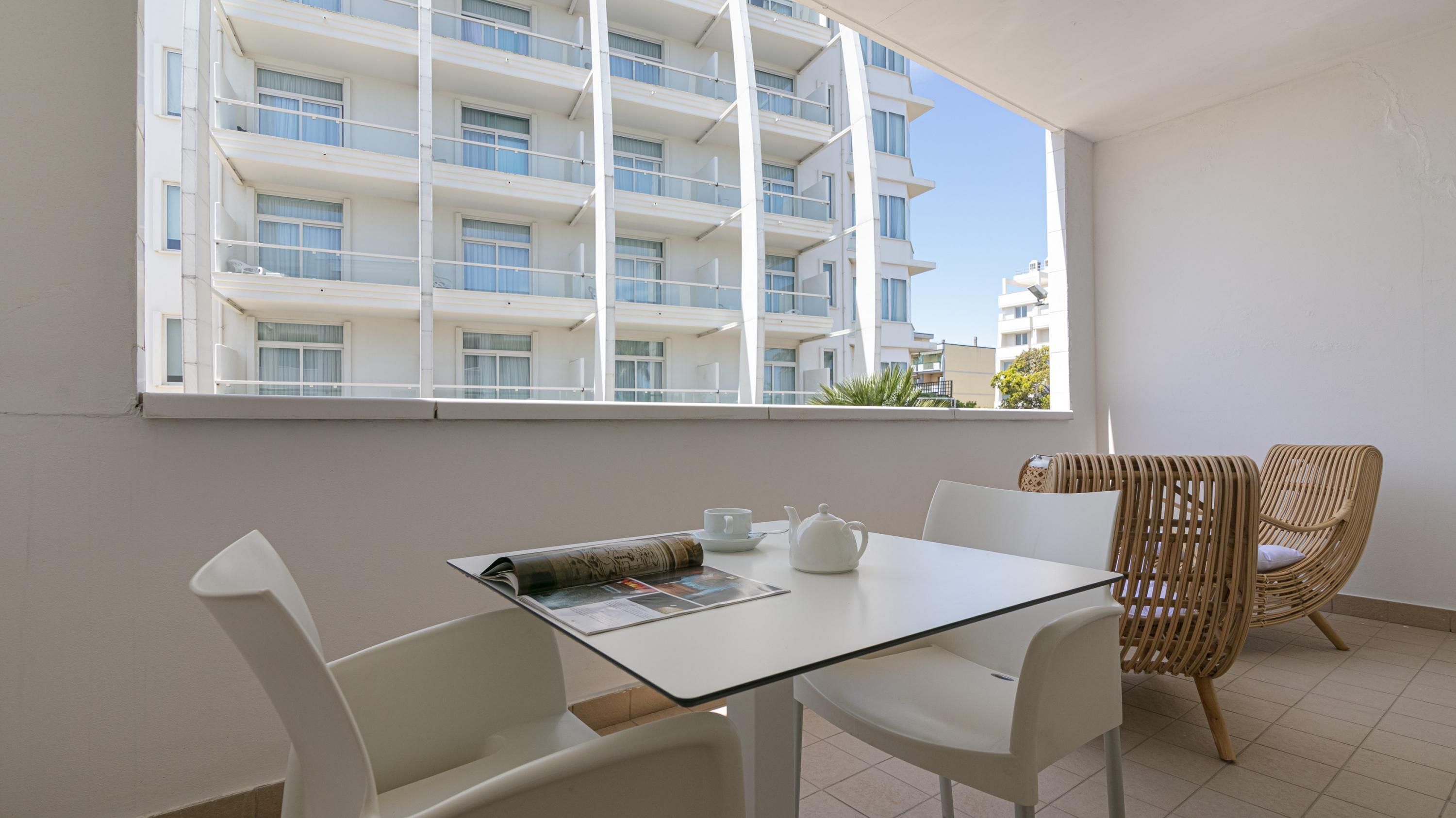 A balcony with a glass table, white chairs, a wicker chair, and a view of a modern building.
