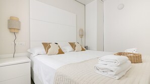 Un lit bien fait, avec une literie blanche et des coussins à motifs, une table de chevet avec une lampe, et un panier en osier contenant des serviettes, le tout posé sur un plancher en bois.