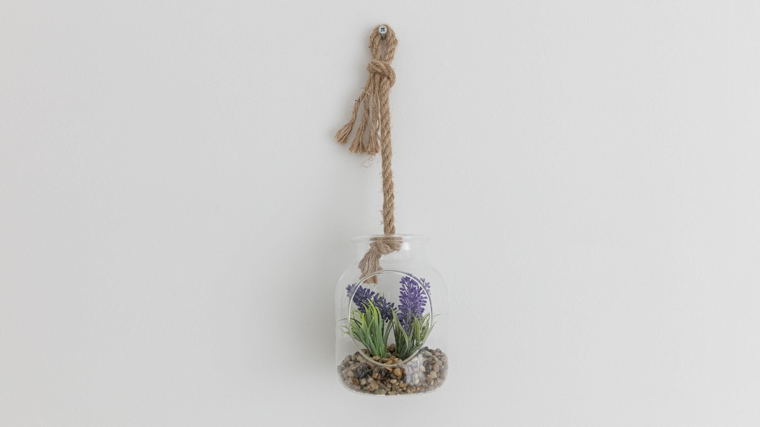 A clear glass jar with a rope handle, containing purple flowers and green foliage, set against a plain background.
