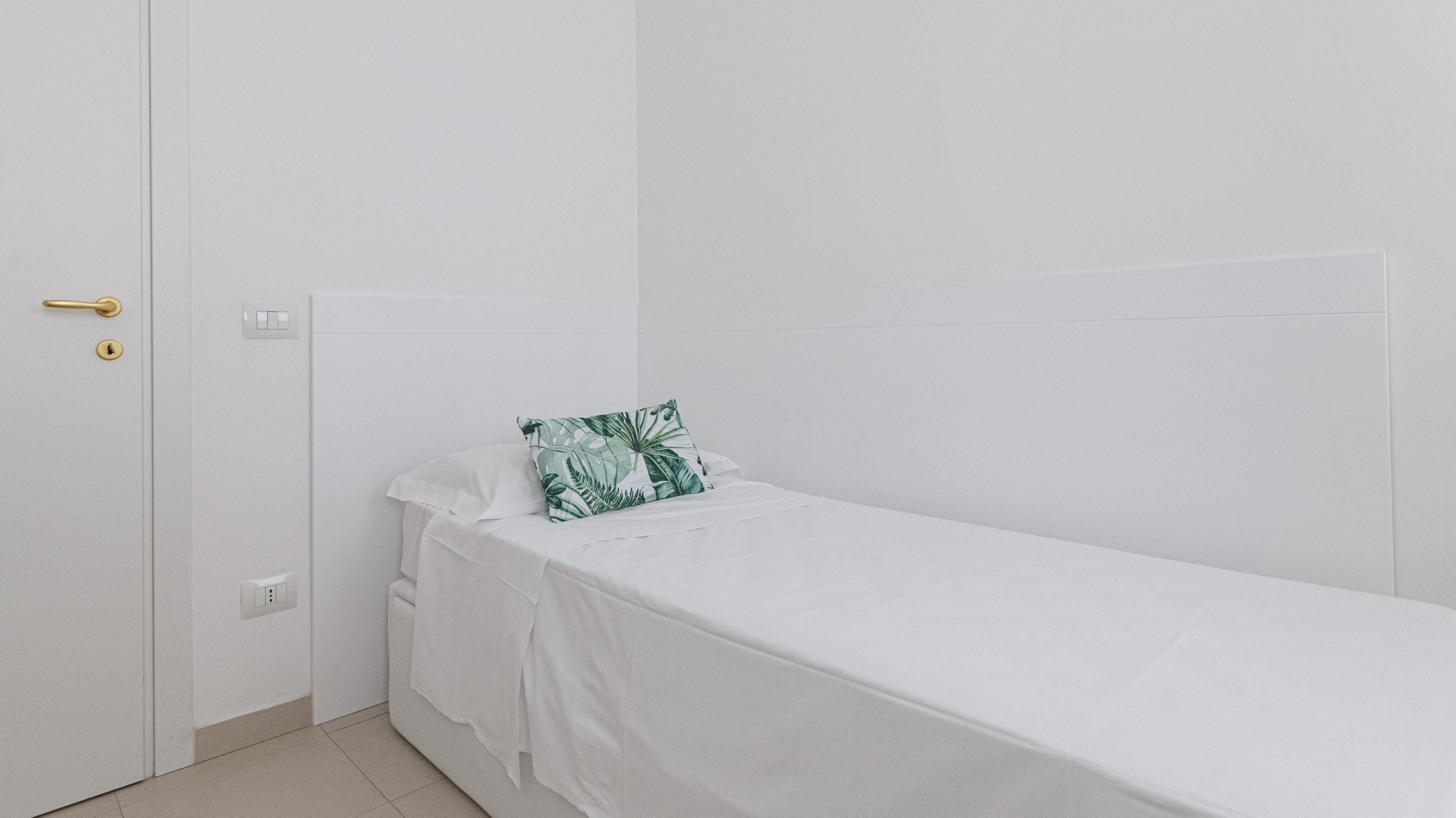 A small, single-bed room with a white bedspread, a green leaf-patterned pillow, a white door with a gold handle, and a white wall.