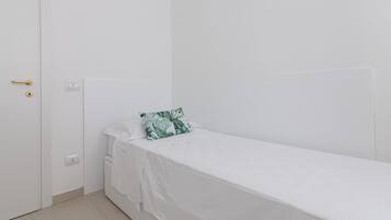 A small, single-bed room with a white bedspread, a green leaf-patterned pillow, a white door with a gold handle, and a white wall.
