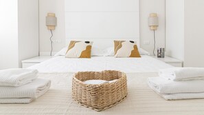 Un lit bien fait, avec une literie blanche et deux coussins décoratifs, un panier en osier posé sur le sol et des serviettes pliées de chaque côté.