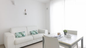 En hvid sofa med to puder, et lille bord med en kop og en plante, samt et vindue med lette gardiner.