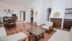 A spacious living room with a wooden table, two white armchairs, and a sofa. There is a kitchen visible through an open doorway, and a fireplace with decorative plates on the wall. - Villa Chiara in Costa Rei (Muravera)