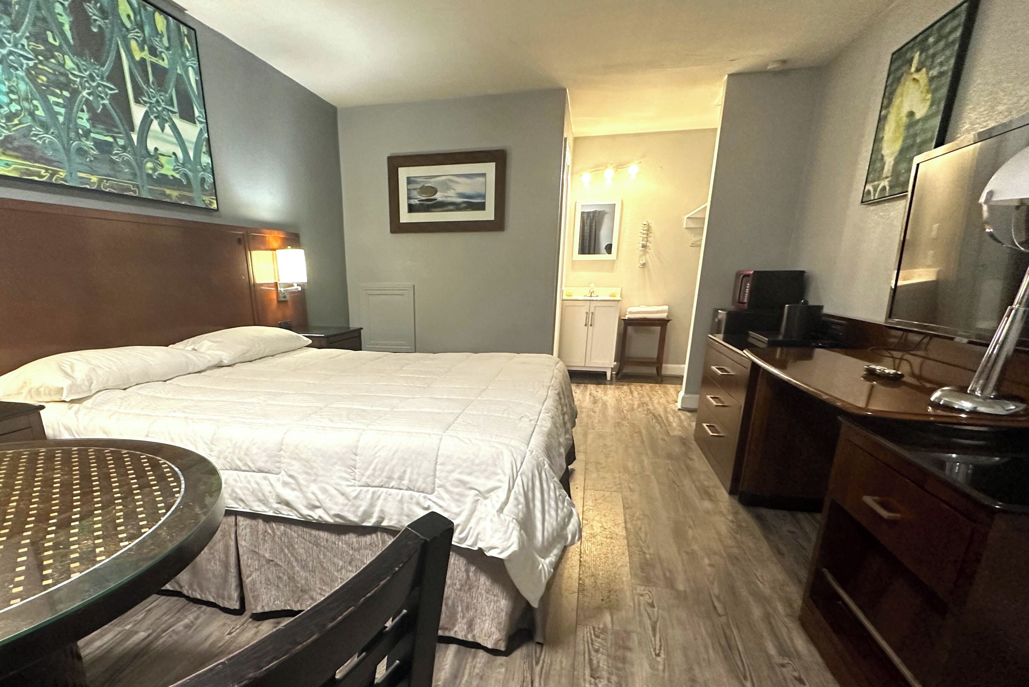 Una camera d'albergo con un letto, una scrivania, una sedia, un tavolino, una cassettiera, uno specchio, un lavandino e un quadro appeso al muro.