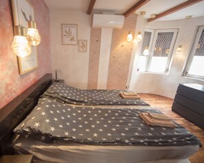 Un dormitorio con una cama que tiene una colcha estampada con motivos de estrellas, un cabecero de madera y una mesita de noche. Hay luminarias fijadas a la pared y una obra de arte enmarcada.