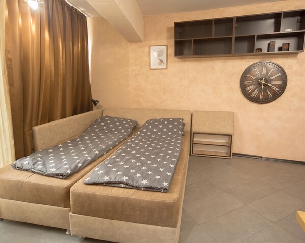 Una habitación con sofá cama, mesita de noche, reloj de pared y estante con objetos decorativos.
