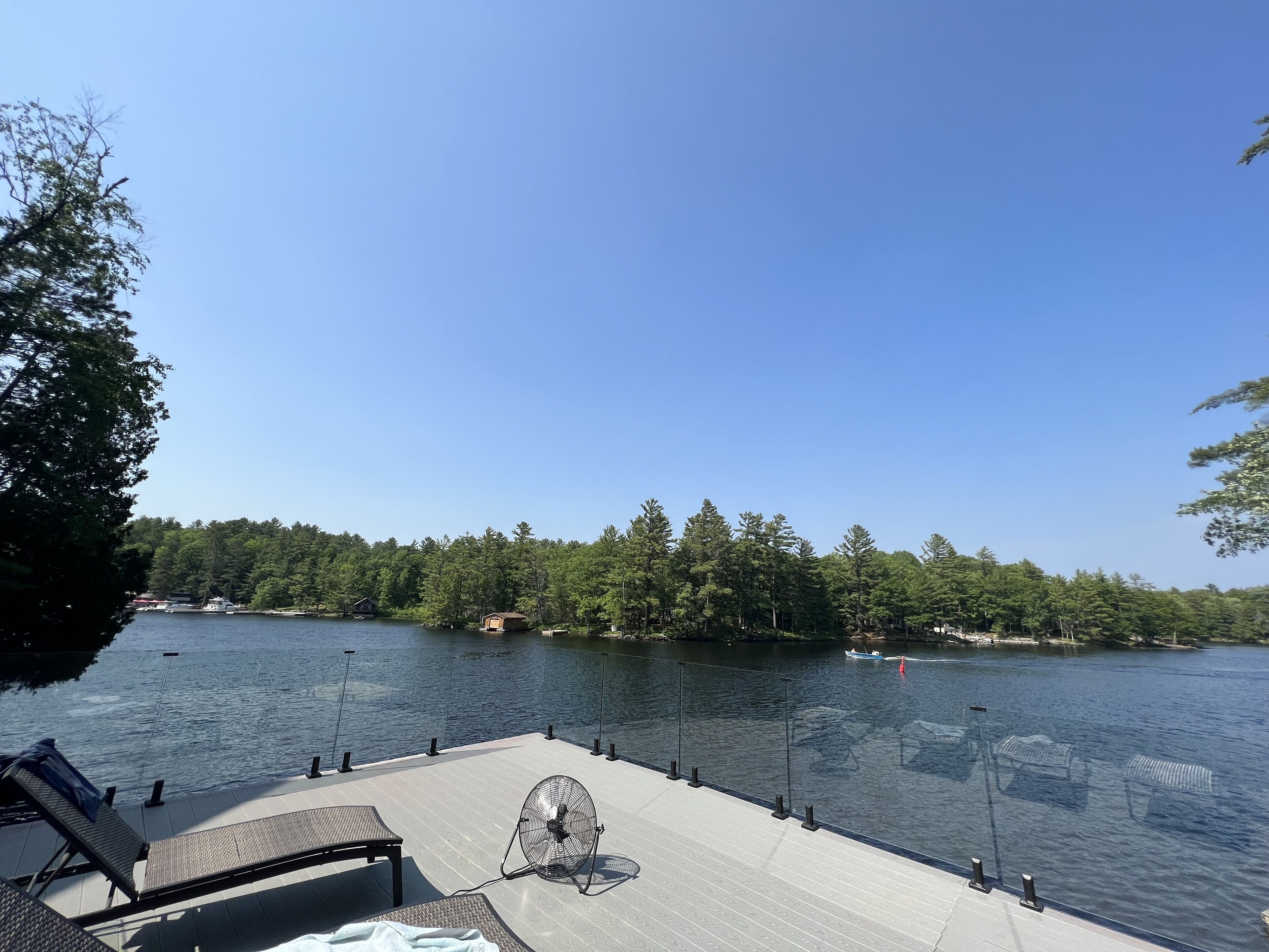A lakeside deck with lounge chairs, a fan, and a view of the lake and forest.