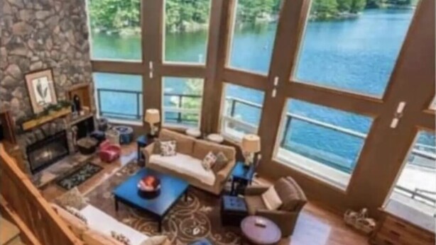 A spacious living area with a stone fireplace, large windows offering a view of a lake, and a cozy seating arrangement. - STUNNING LARGE CHALET COTTAGE IN MUSKOKA (Coldwater)