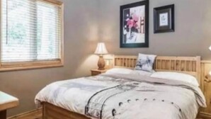 A bedroom with a wooden headboard, a bed with a patterned bedspread, a nightstand with a lamp, and two framed pictures on the wall. - STUNNING LARGE CHALET COTTAGE IN MUSKOKA (Coldwater)