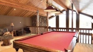 A pool table in a room with wooden beams on the ceiling, stone wall, and a view through a balcony. - STUNNING LARGE CHALET COTTAGE IN MUSKOKA (Coldwater)