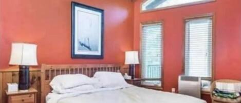 Un dormitorio con techo de madera, una cama con ropa de cama blanca, dos mesitas de noche con lámparas y una ventana con persianas.