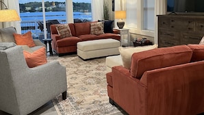 A spacious living room with a large window offering a view of a body of water and hills. There are two sofas, a coffee table, and a TV stand.