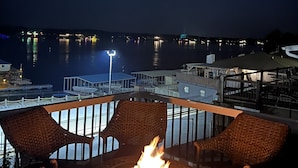 A deck with wicker chairs and a central fire feature, overlooking a body of water with distant city lights.