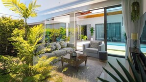 A modern living room with a sofa, armchairs, and a coffee table. There is a large window with a view of a swimming pool and greenery outside.