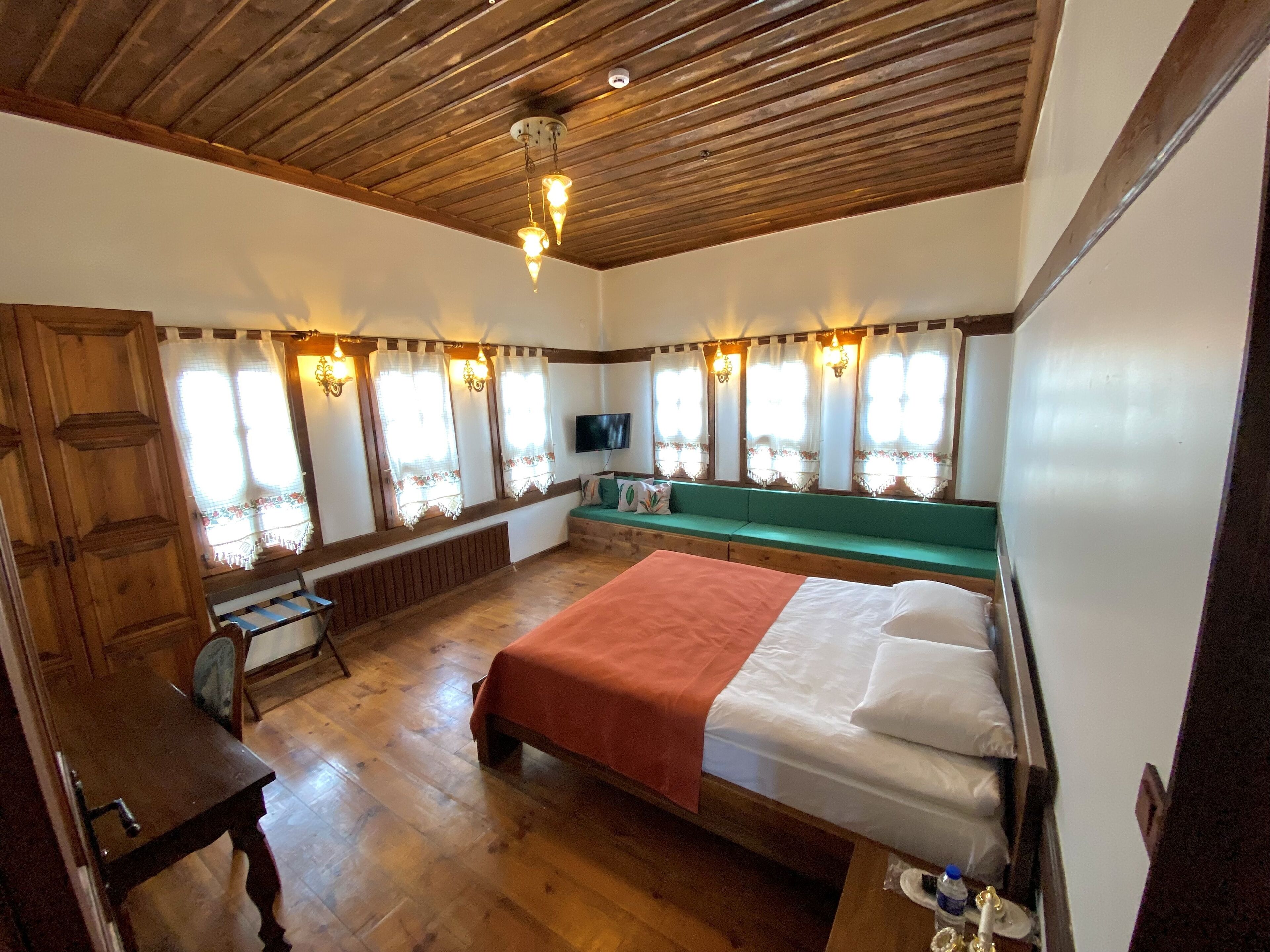 A room with a wooden ceiling, a bed with a red blanket, a bench with cushions, a wooden table, and a television on the wall.