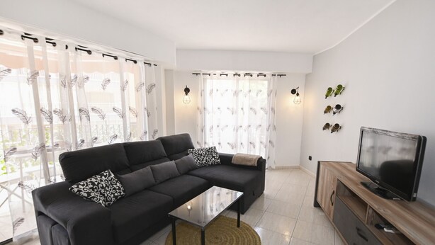 Ein modernes Wohnzimmer mit einem Ecksofa, einem Glastisch, einem Fernseher auf einem Holzständer und einem Balkon mit leichten Vorhängen.