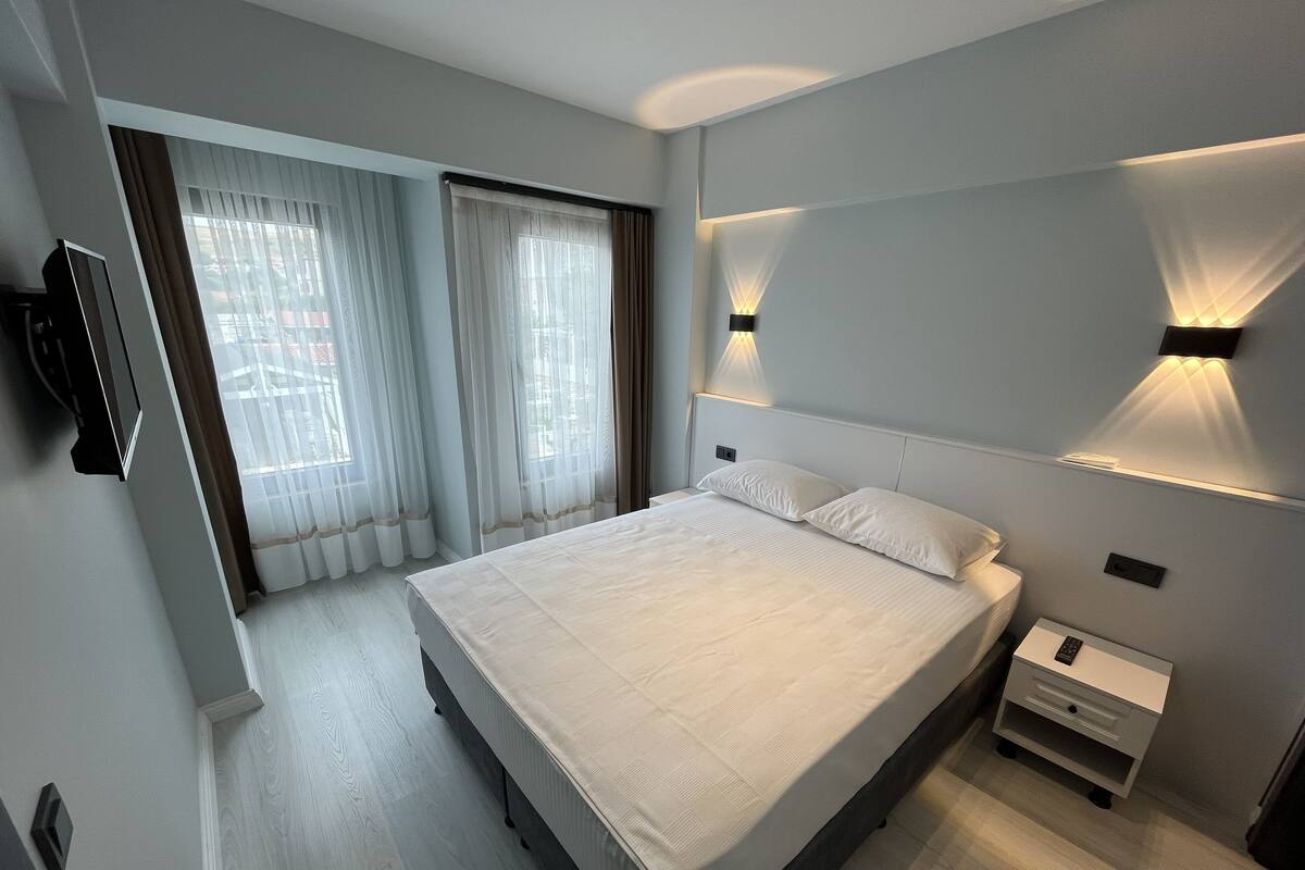 A modern hotel room with a large bed, a bedside table, a TV, and a window with curtains.