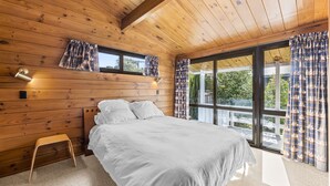 Un dormitorio con techo de madera, cama con ropa de cama blanca y ventanas grandes con cortinas estampadas.