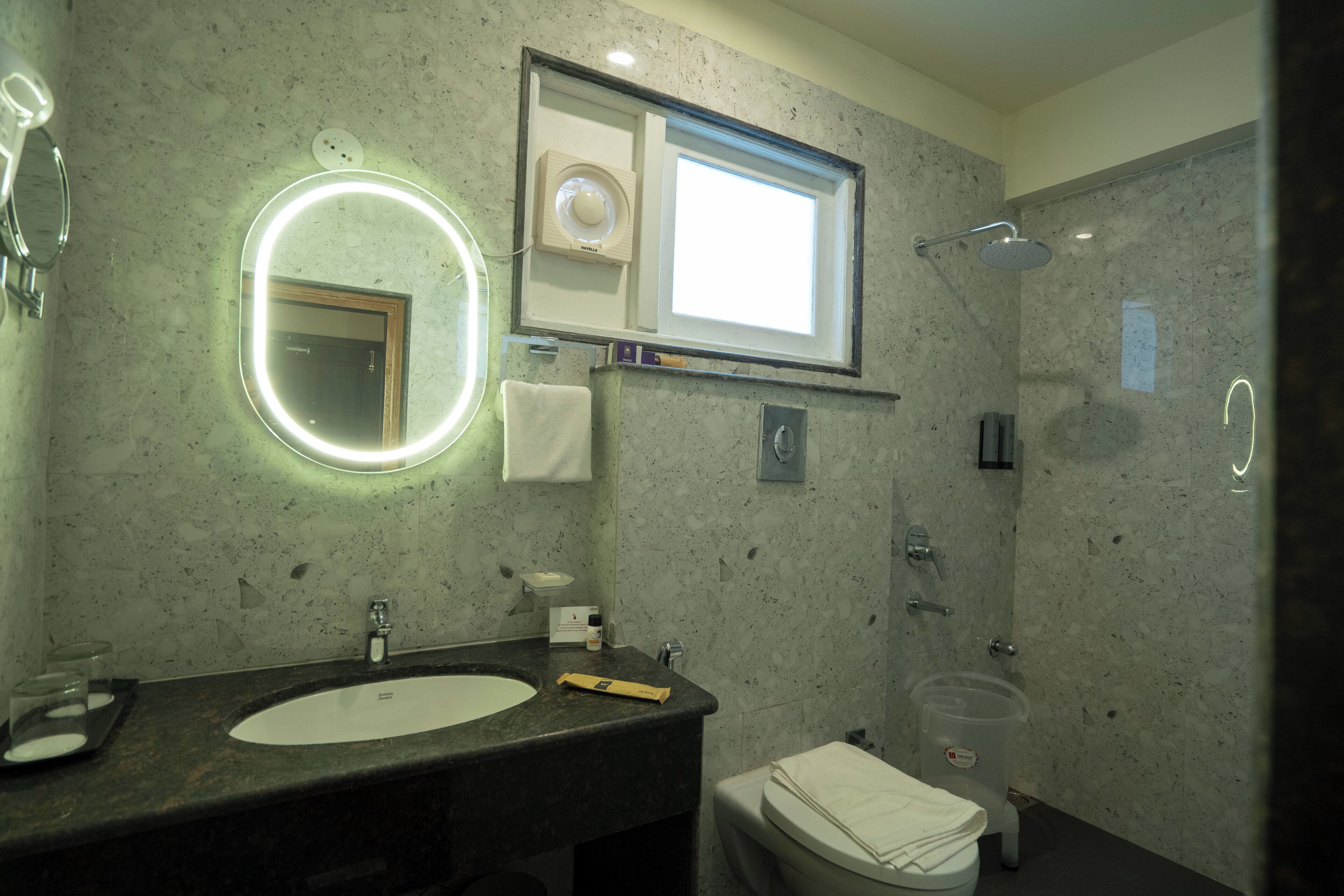 A bathroom with a marble sink, a round mirror with lights, a toilet, and a shower.