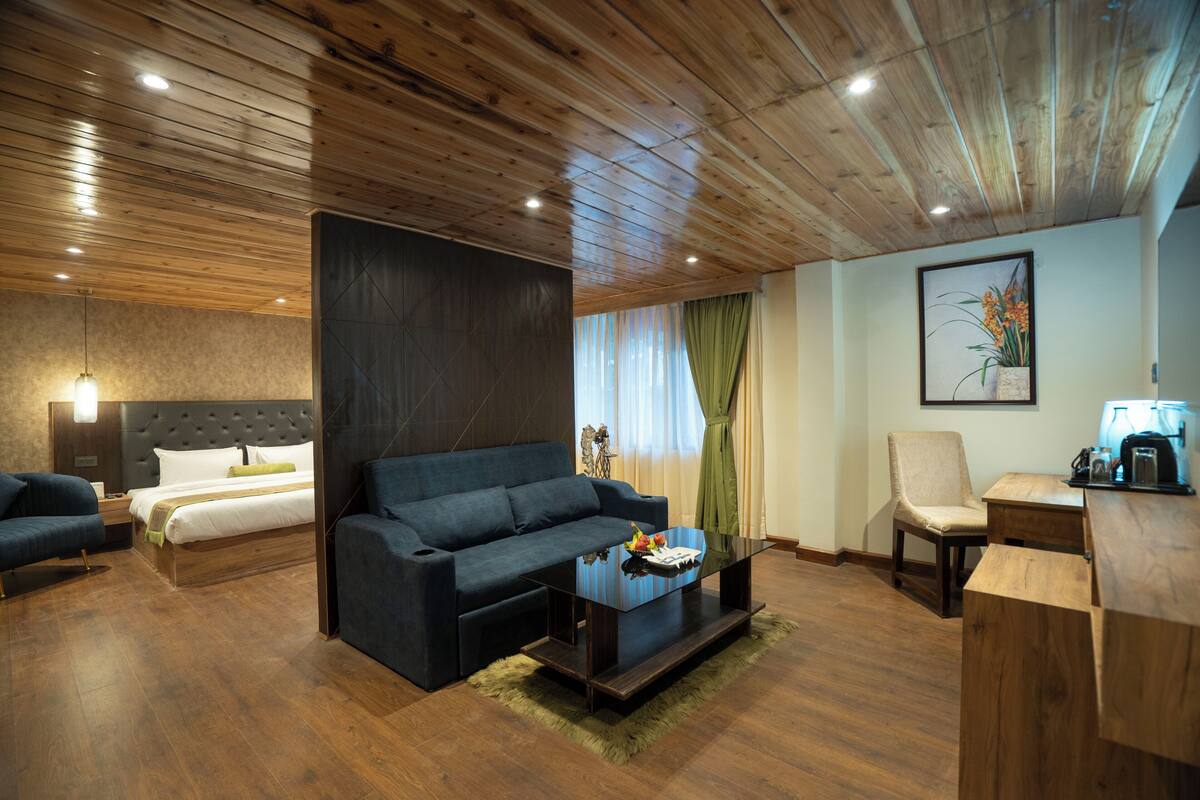 A modern hotel room with a wooden ceiling, a large bed, a dark sofa, a coffee table, and a desk with a lamp.
